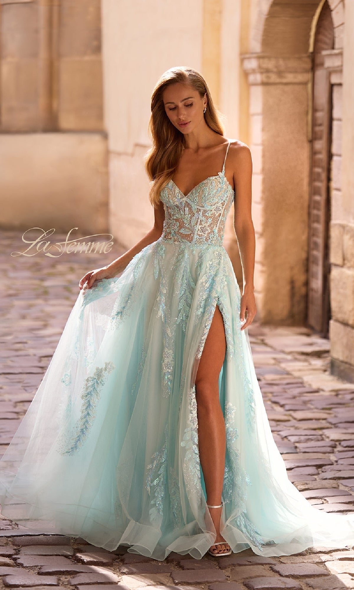 A woman stands on a cobblestone street wearing the La Femme 32810 long prom dress, featuring light blue tulle, beaded lace applique, thin straps, and a high slit.
